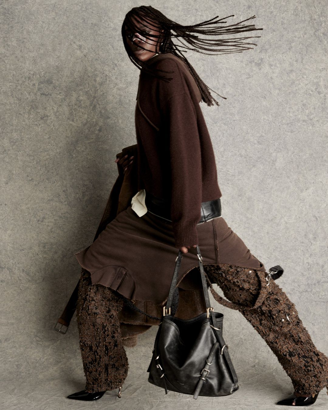 A stylish adult female stands in a studio wearing a brown knit hoodie, skirt, heeled boots, and carrying a black leather shoulder bag.