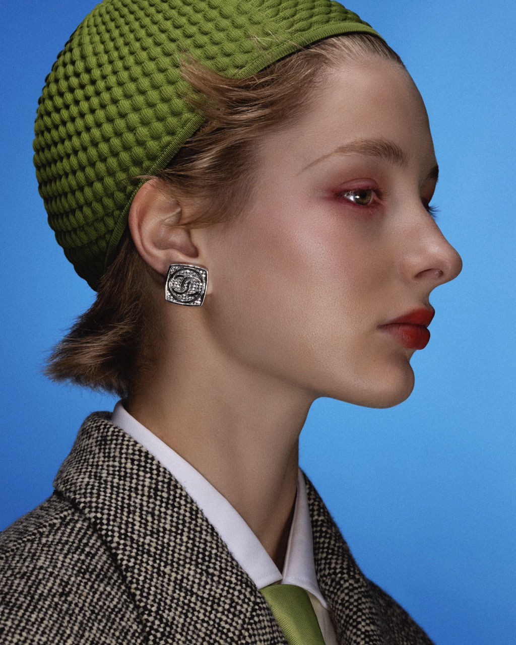 Profile of a woman in a textured green hat with silver earrings against a bright blue background, wearing a tweed jacket.