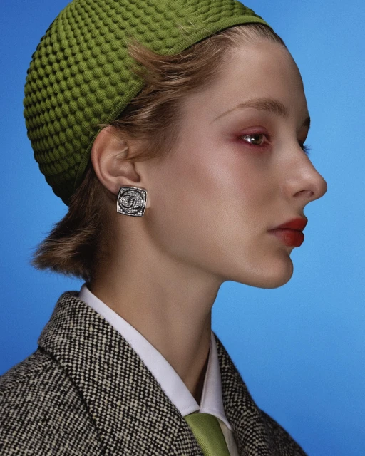Profile of a woman in a textured green hat with silver earrings against a bright blue background, wearing a tweed jacket.
