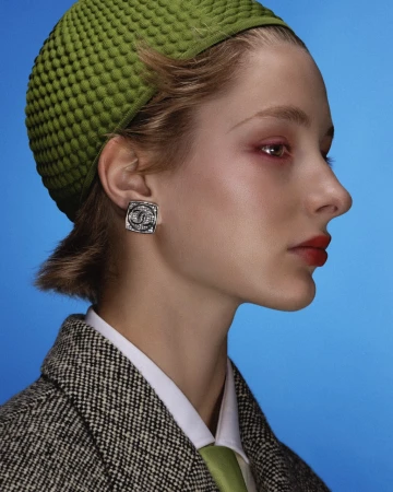 Profile of a woman in a textured green hat with silver earrings against a bright blue background, wearing a tweed jacket.