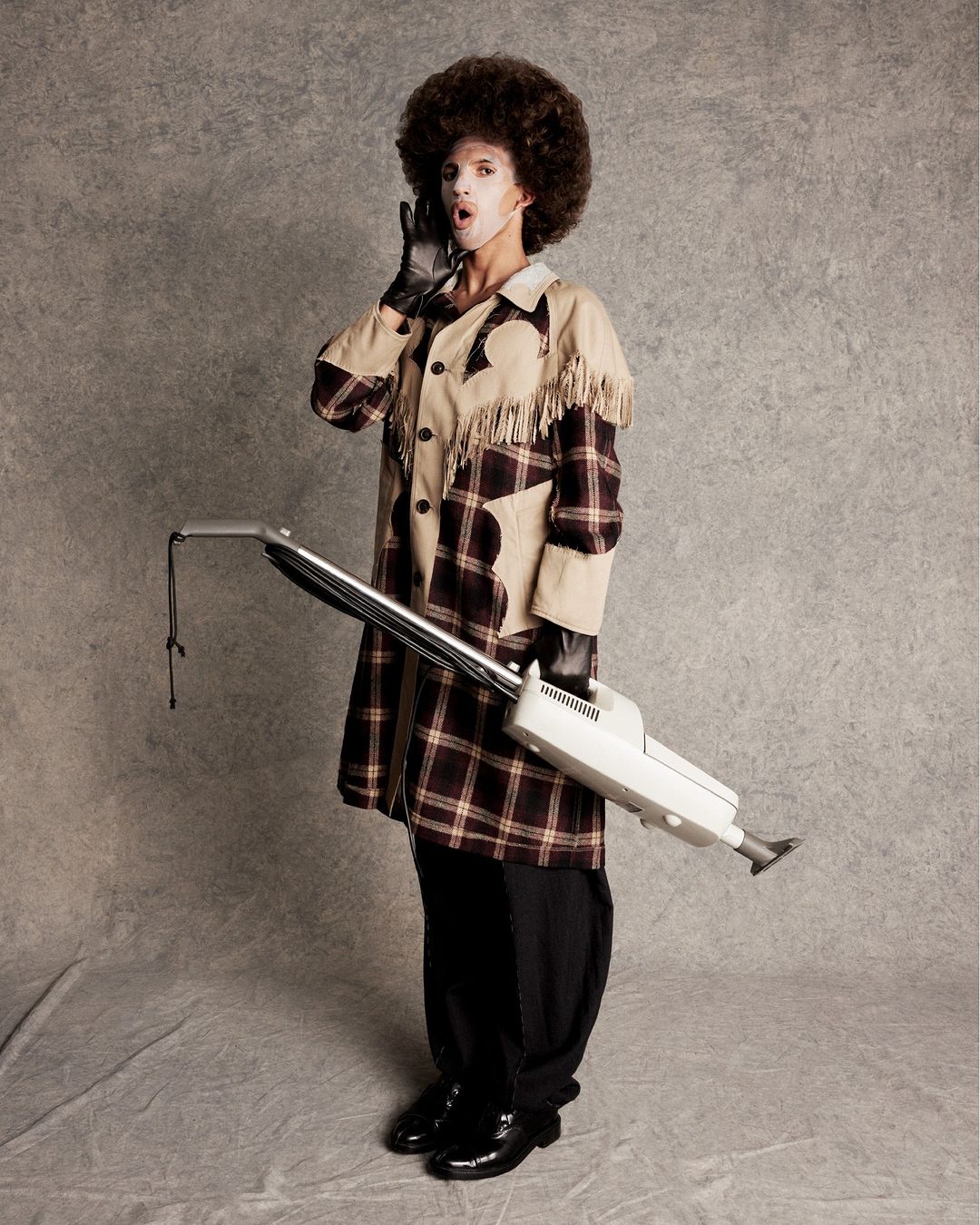 A person in a plaid coat, black gloves, and afro wig is holding a vacuum cleaner and cupping their hand to their mouth in a studio shot.