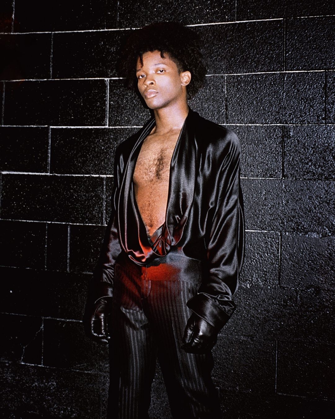Portrait of an androgynous person with afro hair posing in a black silk outfit against a black brick wall in a dark indoor environment.