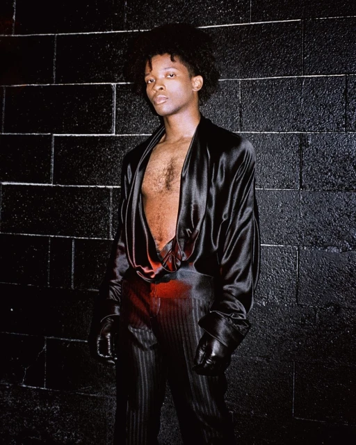 Portrait of an androgynous person with afro hair posing in a black silk outfit against a black brick wall in a dark indoor environment.
