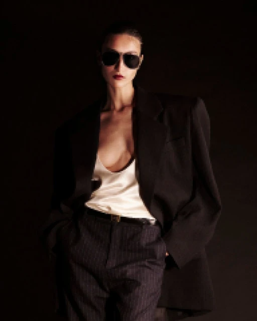 Fashion portrait of an adult woman in a blazer, white top, and pinstripe trousers with sunglasses, set against a dark background.