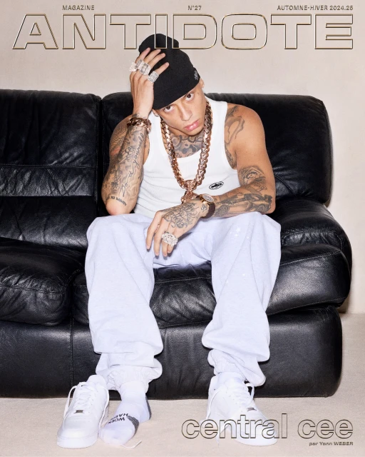 Central Cee sits on a black leather couch in a magazine cover photo, wearing a white tank top, grey sweatpants, jewelry, and sneakers.