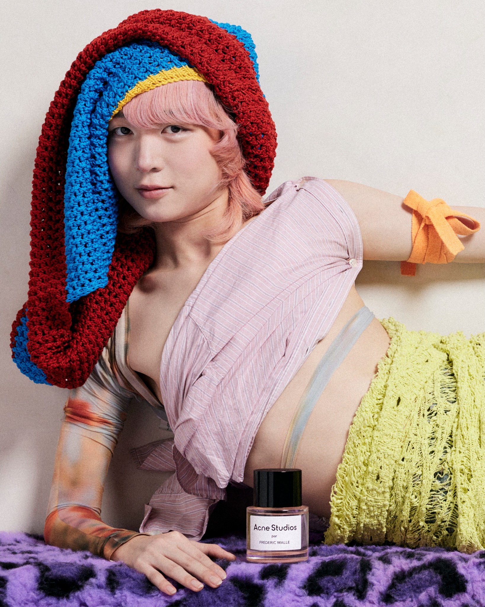 Person with pink hair and a crochet hat poses next to an Acne Studios perfume bottle on a purple fur blanket in a studio.