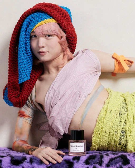 Person with pink hair and a crochet hat poses next to an Acne Studios perfume bottle on a purple fur blanket in a studio.