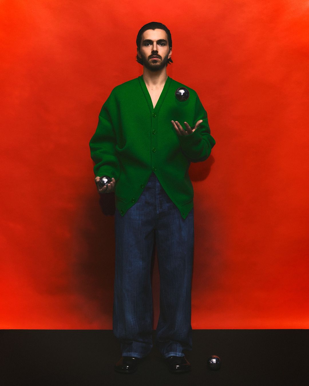 Man in a green cardigan stands against a red wall juggling silver spheres. He wears dark jeans and black shoes on a black floor.
