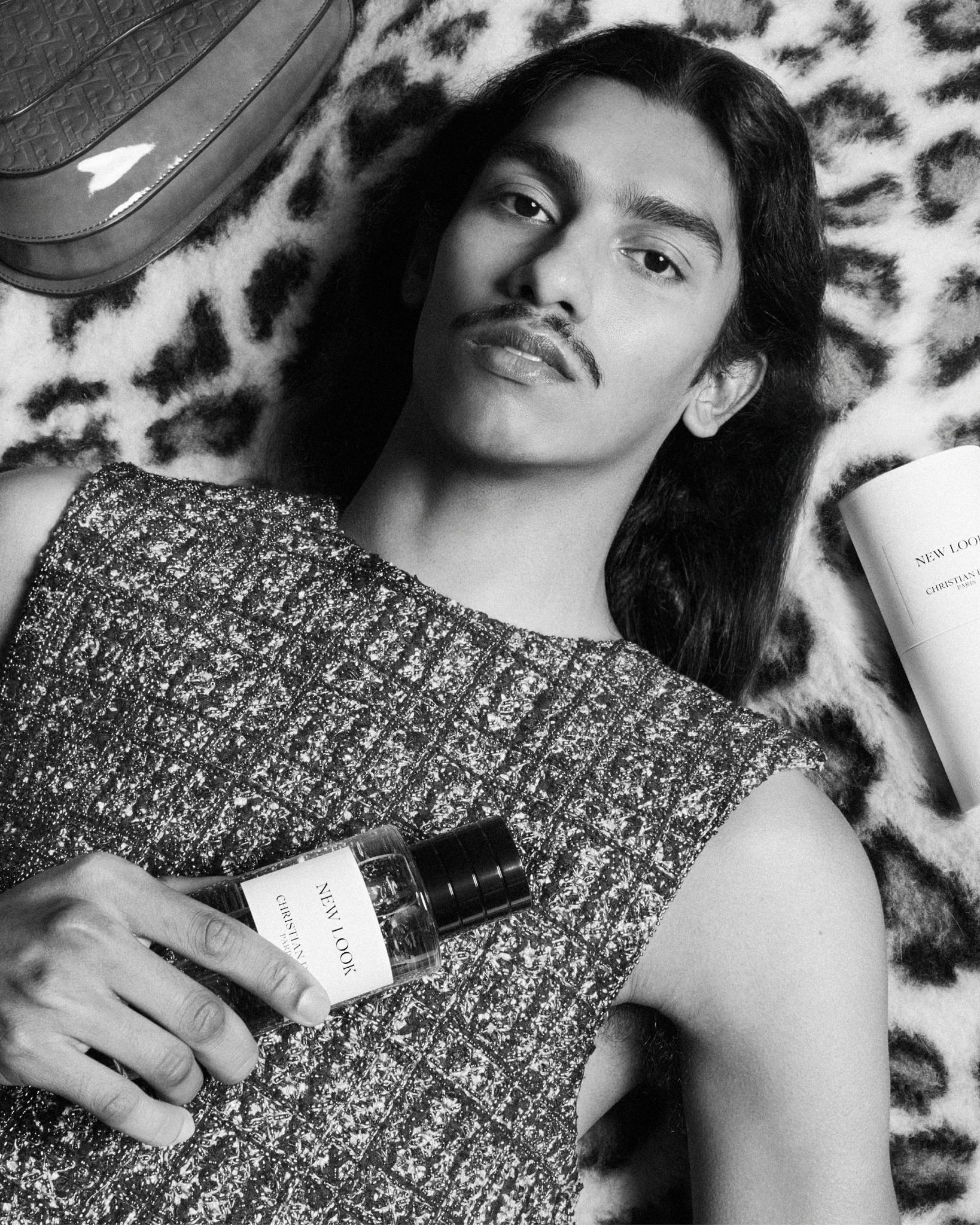Portrait of a slender adult male with long dark hair and a mustache holding a Dior New Look perfume bottle while lying on an animal print.