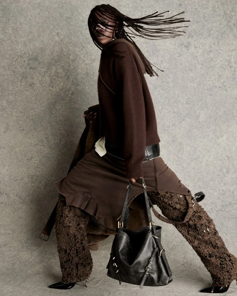 Model with long braided hair wearing brown knitwear and textured trousers holding a black leather handbag