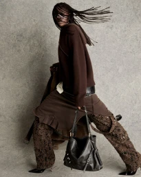 Model with long braided hair wearing brown knitwear and textured trousers holding a black leather handbag