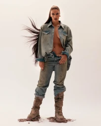 Woman in an unbuttoned denim shirt and mud-stained jeans wearing heavy boots, with her long hair blowing.