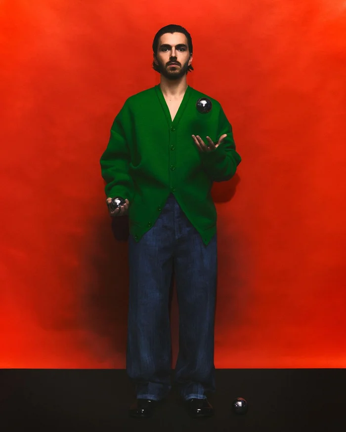 Man wearing a green cardigan and denim jeans juggling chrome spheres against a vibrant red backdrop