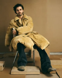 Man with dark hair and beard wearing an oversized tan coat and dark trousers seated on a chair in a studio.