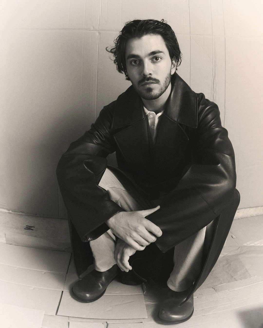 A man wearing a black leather coat squatting indoors with a plain backdrop, photographed by Yann Weber in Paris.
