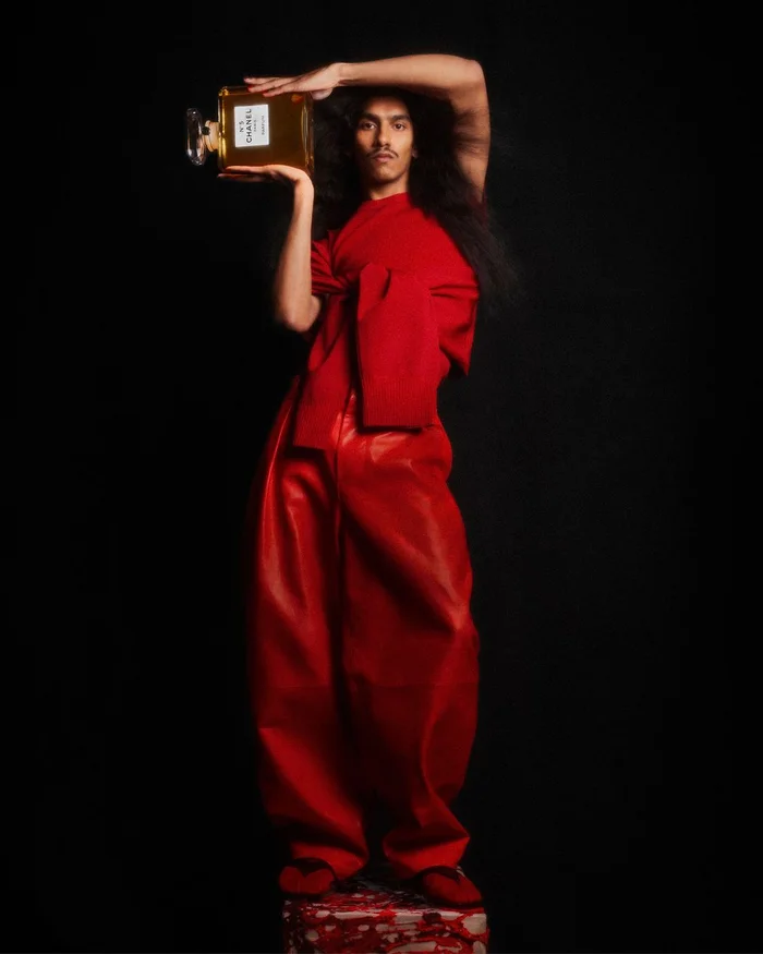 Man with long hair wearing red clothing lifting a large Chanel perfume bottle above his head against a dark studio background