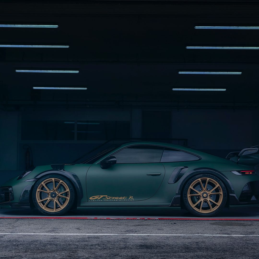 Side view of a matte-green Porsche GT Street R parked indoors with gold wheels and black trim, in a dimly lit automotive setting.