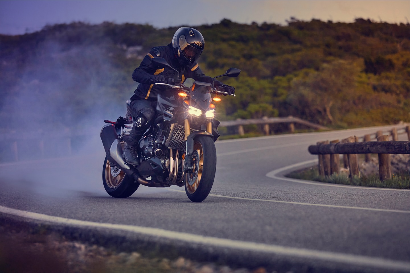 Man riding a black motorcycle on a winding road with green hills at dusk, creating a dynamic image of speed and adventure.