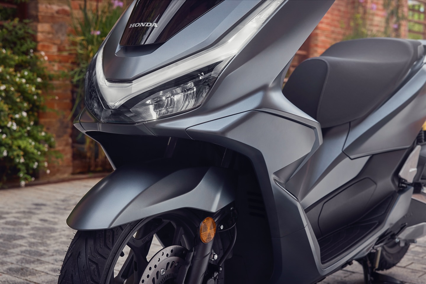 Close-up of a gray Honda PCX scooter on a paved surface, highlighting its modern design and sleek lines against a blurred brick wall background.