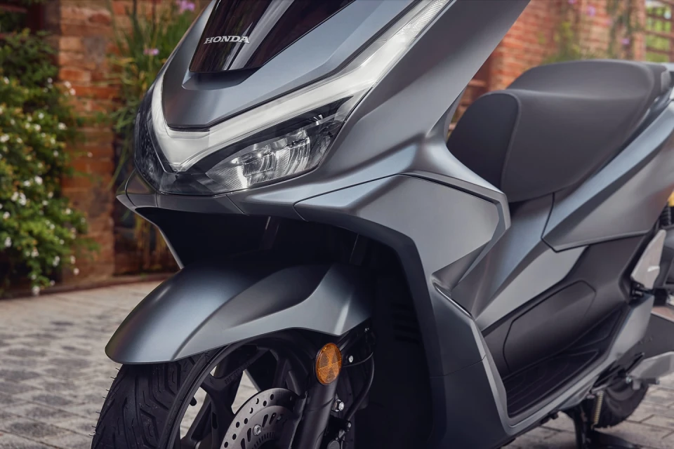 Close-up of a gray Honda PCX scooter on a paved surface, highlighting its modern design and sleek lines against a blurred brick wall background.