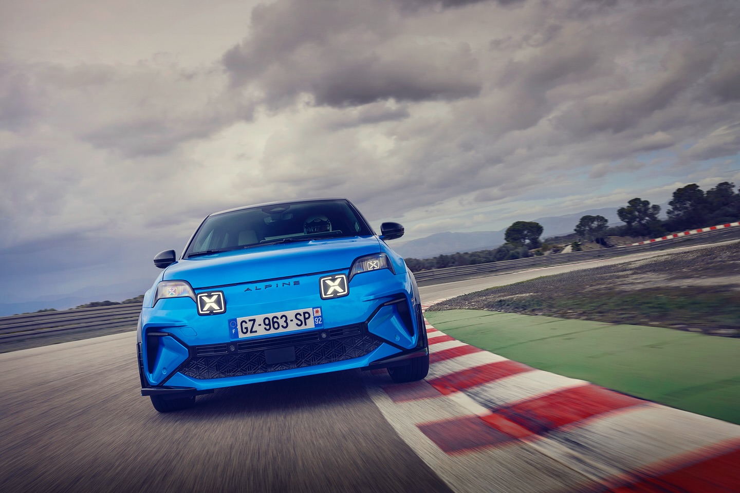 Bright blue Alpine sports car drives on a race track. Modern design with unique headlights. Cloudy sky with visible mountain range.