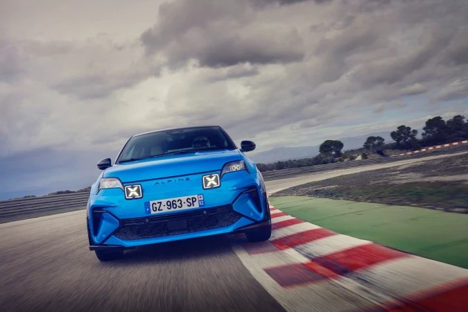 Bright blue Alpine sports car drives on a race track. Modern design with unique headlights. Cloudy sky with visible mountain range.