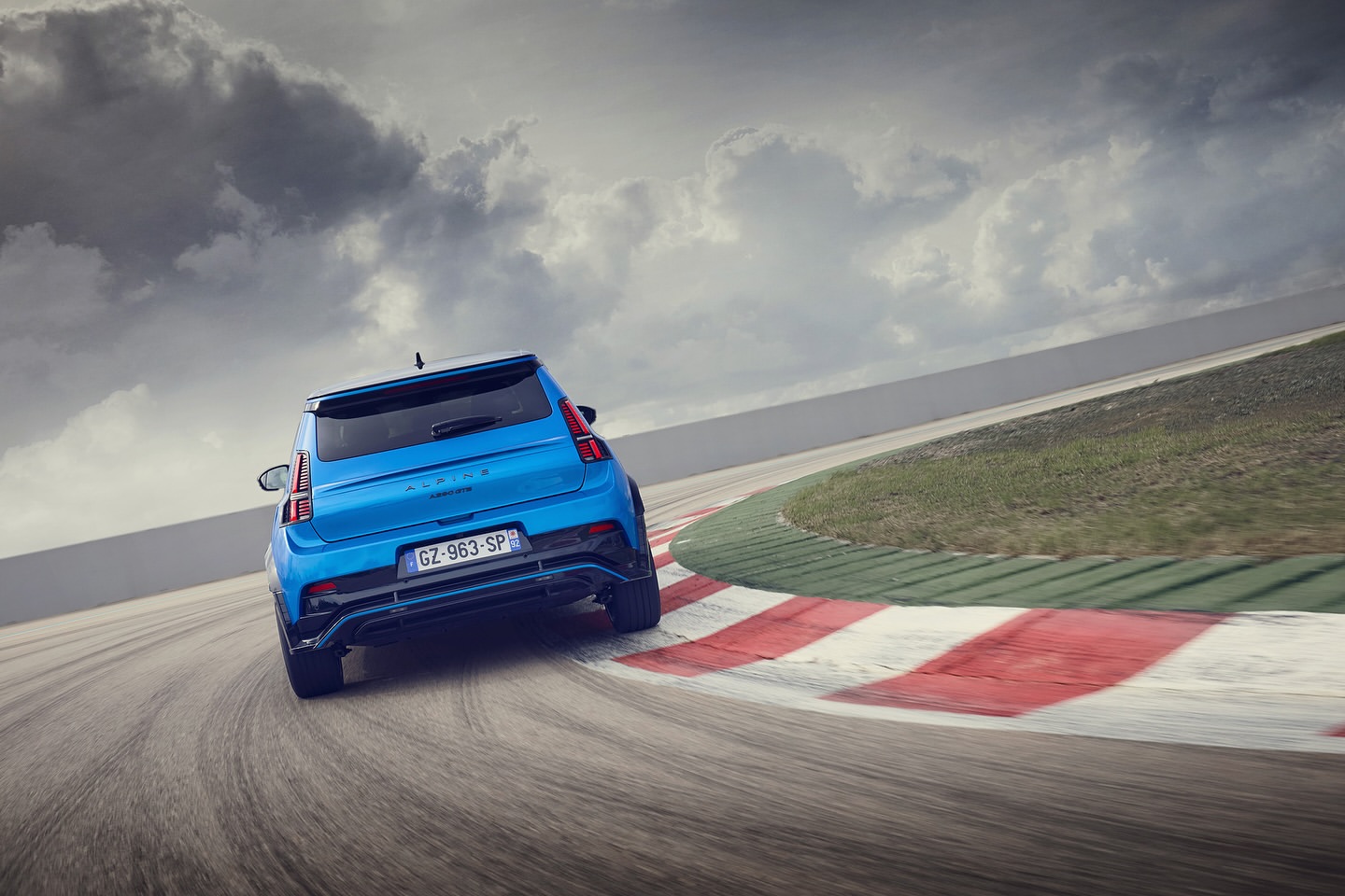 A blue Alpine A110 sports car takes a sharp turn on a race track under a cloudy sky, showcasing its agile handling and sleek design.