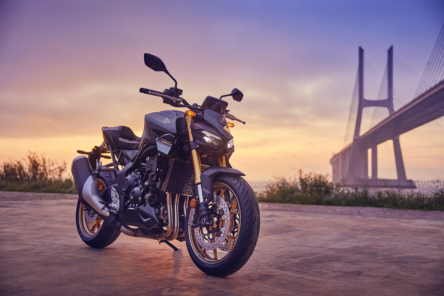 A black and bronze Honda motorcycle parked outdoors on pavement with a large bridge spanning water in the background at sunset.