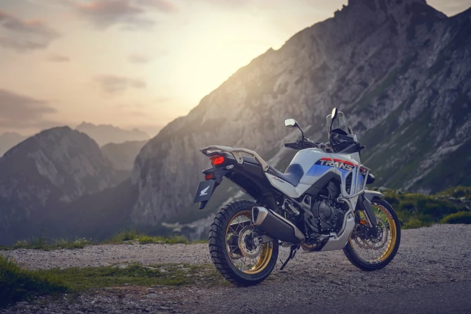 Motorcycle parked on a hilltop road with a mountain range in the background during the golden hour with a sky filled with clouds.