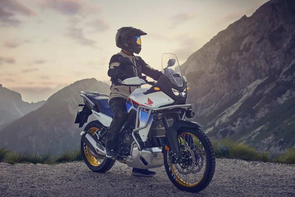 A motorcyclist wearing a helmet sits atop a white, blue, and red touring motorcycle on a gravel path with mountainous background.