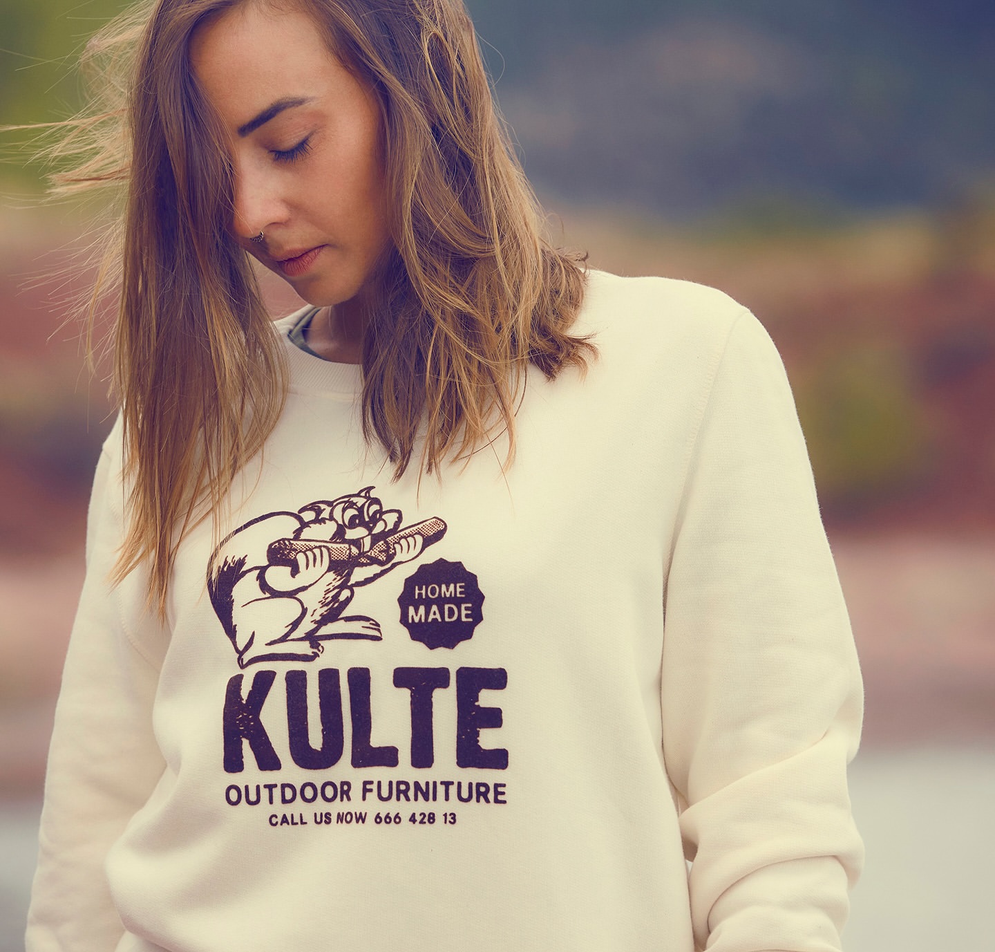 An adult female model poses outdoors wearing a cream-colored crewneck sweatshirt with a beaver illustration and 'Kulte Outdoor Furniture' text.