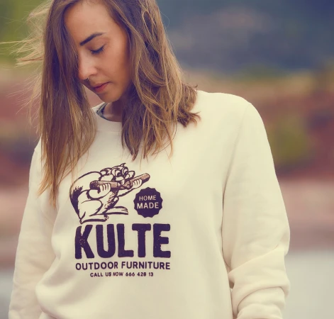An adult female model poses outdoors wearing a cream-colored crewneck sweatshirt with a beaver illustration and 'Kulte Outdoor Furniture' text.