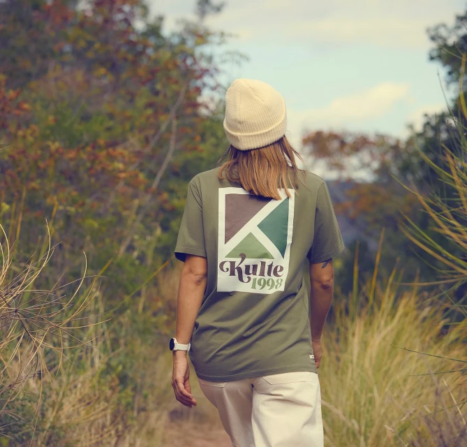 Woman in olive-green shirt and cream-colored beanie stands in a leafy outdoor setting with branded graphic design on the shirt.