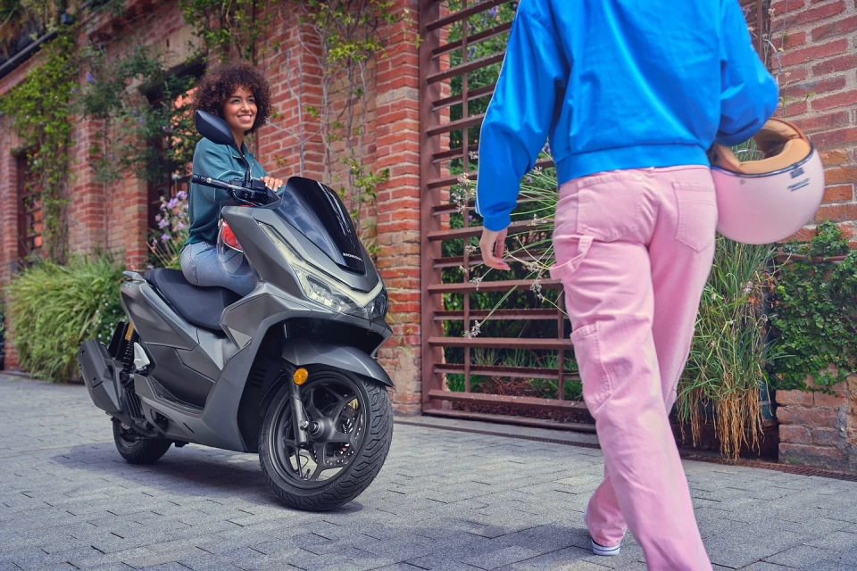 A woman sits on a gray scooter on a brick sidewalk with a second woman walking by holding a helmet in an outdoor setting.