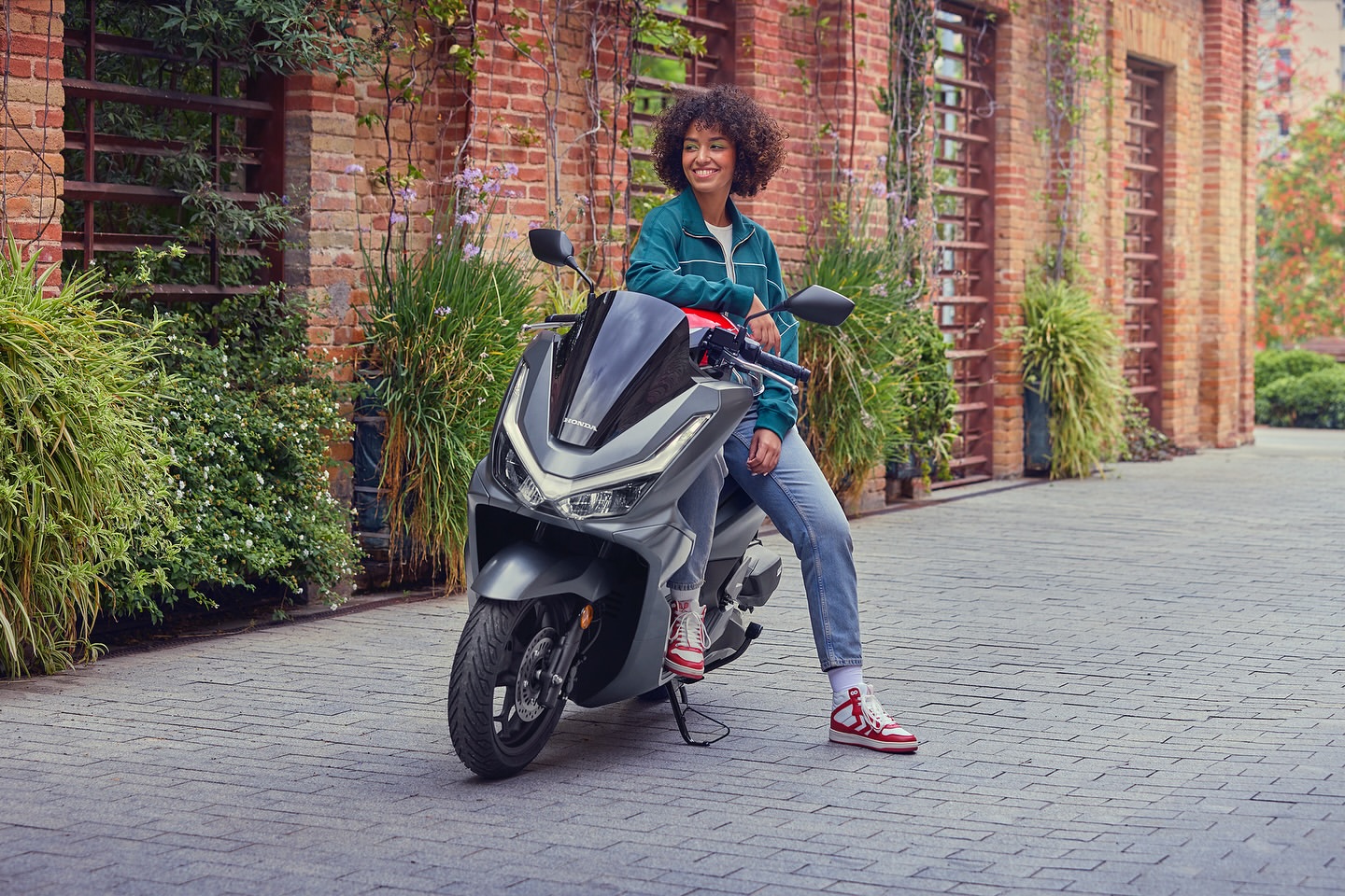 An adult female smiles while sitting on a gray scooter in an outdoor, urban setting with a brick building and greenery visible in the background.