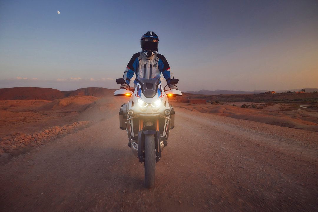 Person riding a motorcycle on a dirt road through a desert landscape at dusk with mountains and a partial moon in the background.