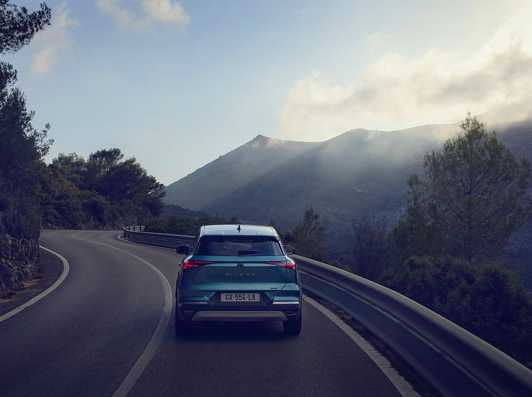 Teal SUV driving on winding road through mountains with trees and blue sky with light clouds, rear view of car.
