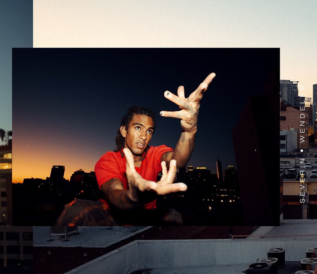 Adult male poses with hands raised against a dark cityscape at twilight. Man with locs in red t-shirt on rooftop with dusk sky backdrop.