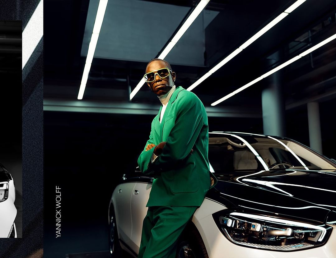 Adult male in green suit stands confidently against a white car in a dark interior with bright overhead lighting, captured by Yannick Wolff.