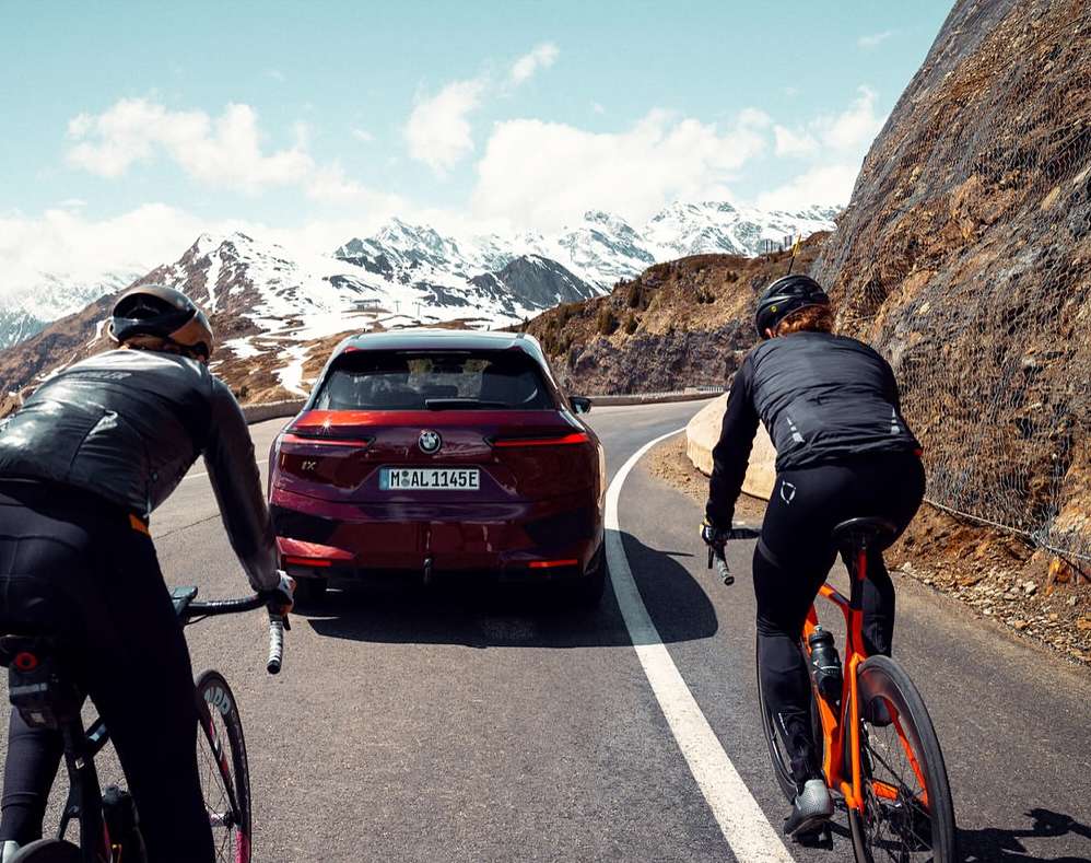 A modern purple BMW iX follows two cyclists wearing helmets on a mountain road with snow-capped mountains in the background under a sunny sky.