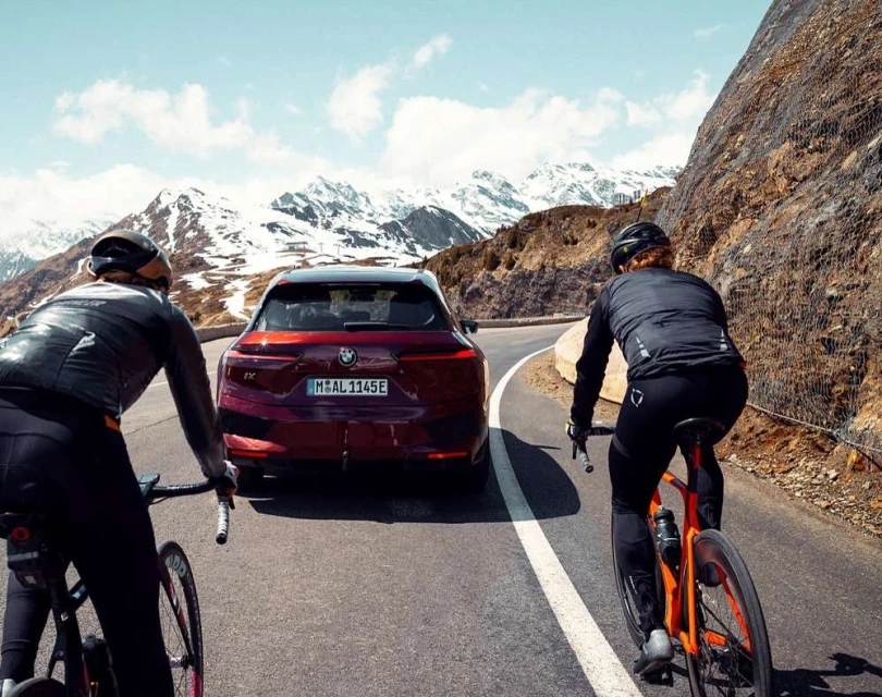 A modern purple BMW iX follows two cyclists wearing helmets on a mountain road with snow-capped mountains in the background under a sunny sky.