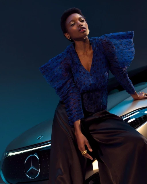 Elegant woman in a blue top and black skirt poses next to a silver Mercedes-Benz EQE car in a studio setting, showcasing automotive fashion.