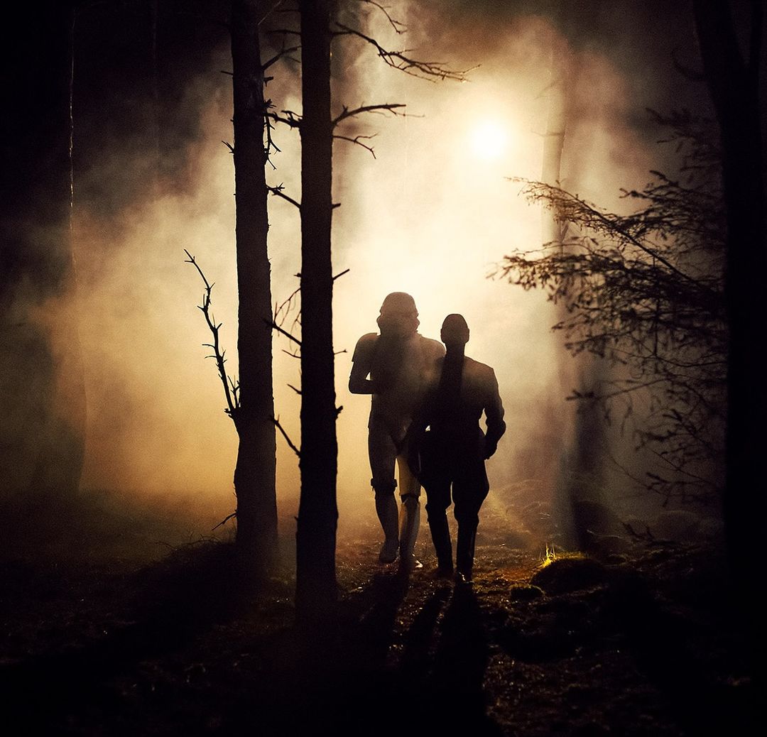 Two figures, one in white armor, the other in a darker outfit, stand in a foggy forest with trees silhouetted against a bright light.