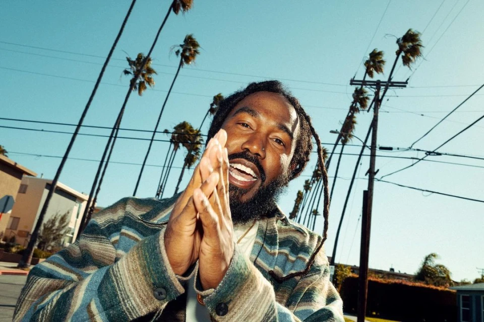 Man with dreadlocks smiles, clasped hands up toward the camera. Palm trees line a street under a clear sky in an urban setting.