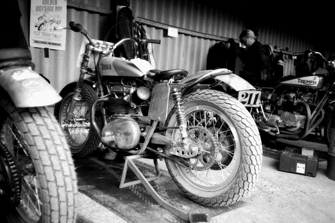 Black and white image of a BSA motorcycle parked indoors, showcasing its engine, wheels, and vintage design, alongside other bikes.