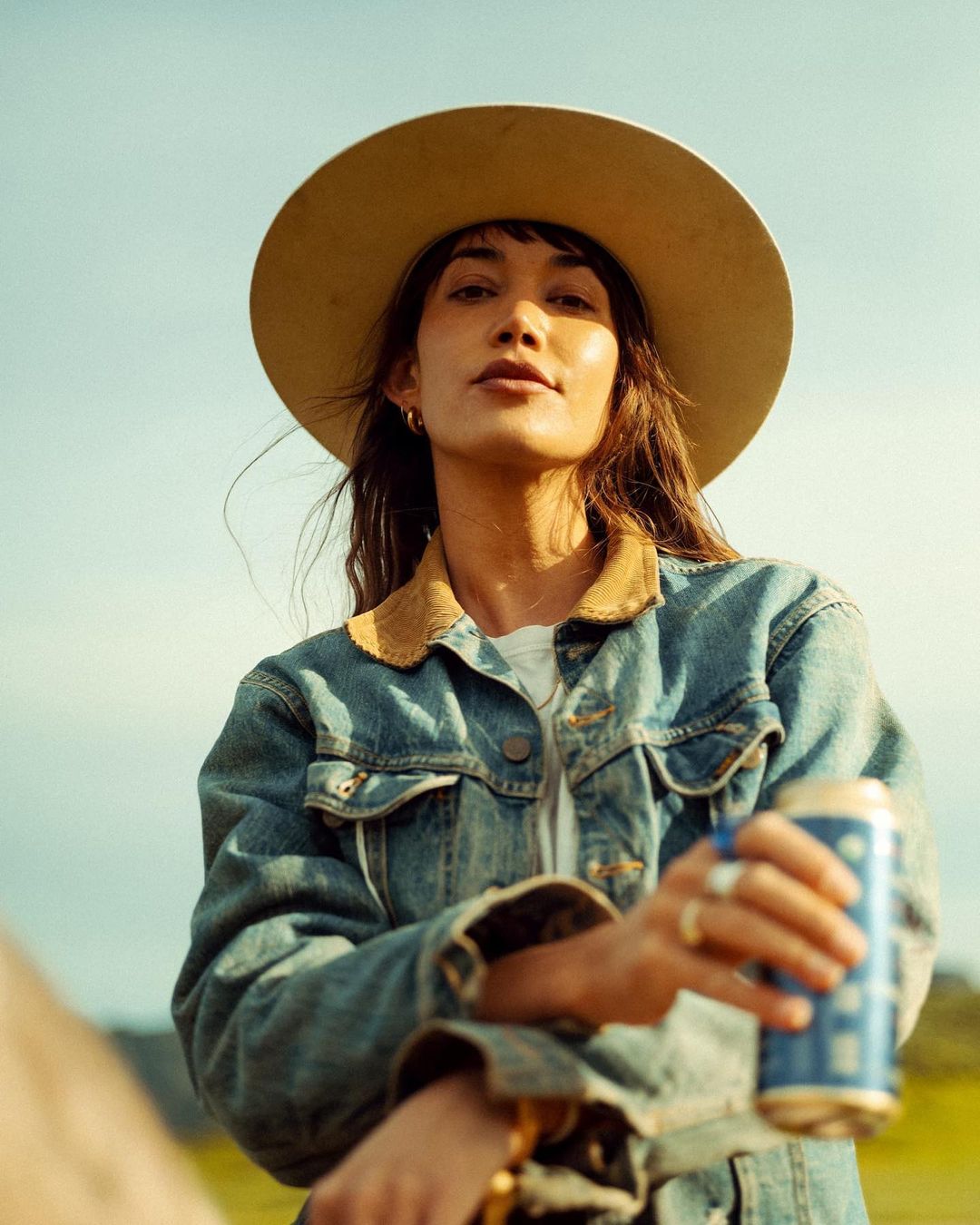 Portrait of a young adult woman with long dark hair wearing a wide brim hat and a denim jacket, holding a blue beer can outdoors.