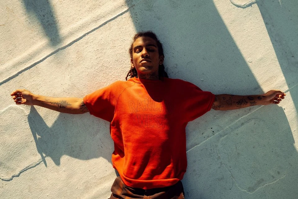 Adult male with dreadlocks in a red sweatshirt lying on a concrete surface with arms outstretched in an outdoor setting, bathed in sunlight.
