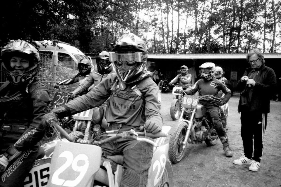 Group of motocross riders on dirt bikes wearing helmets and racing gear preparing on a dirt track, with a coach on the sidelines.