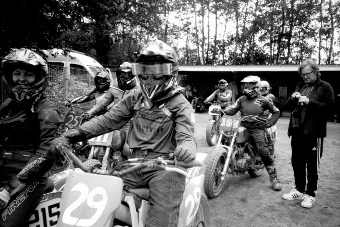 Group of motocross riders on dirt bikes wearing helmets and racing gear preparing on a dirt track, with a coach on the sidelines.