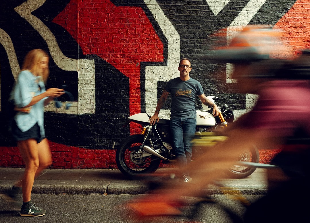 Man poses next to Triumph motorcycle on urban street in front of brick wall with graffiti, while blurred figures cross foreground.